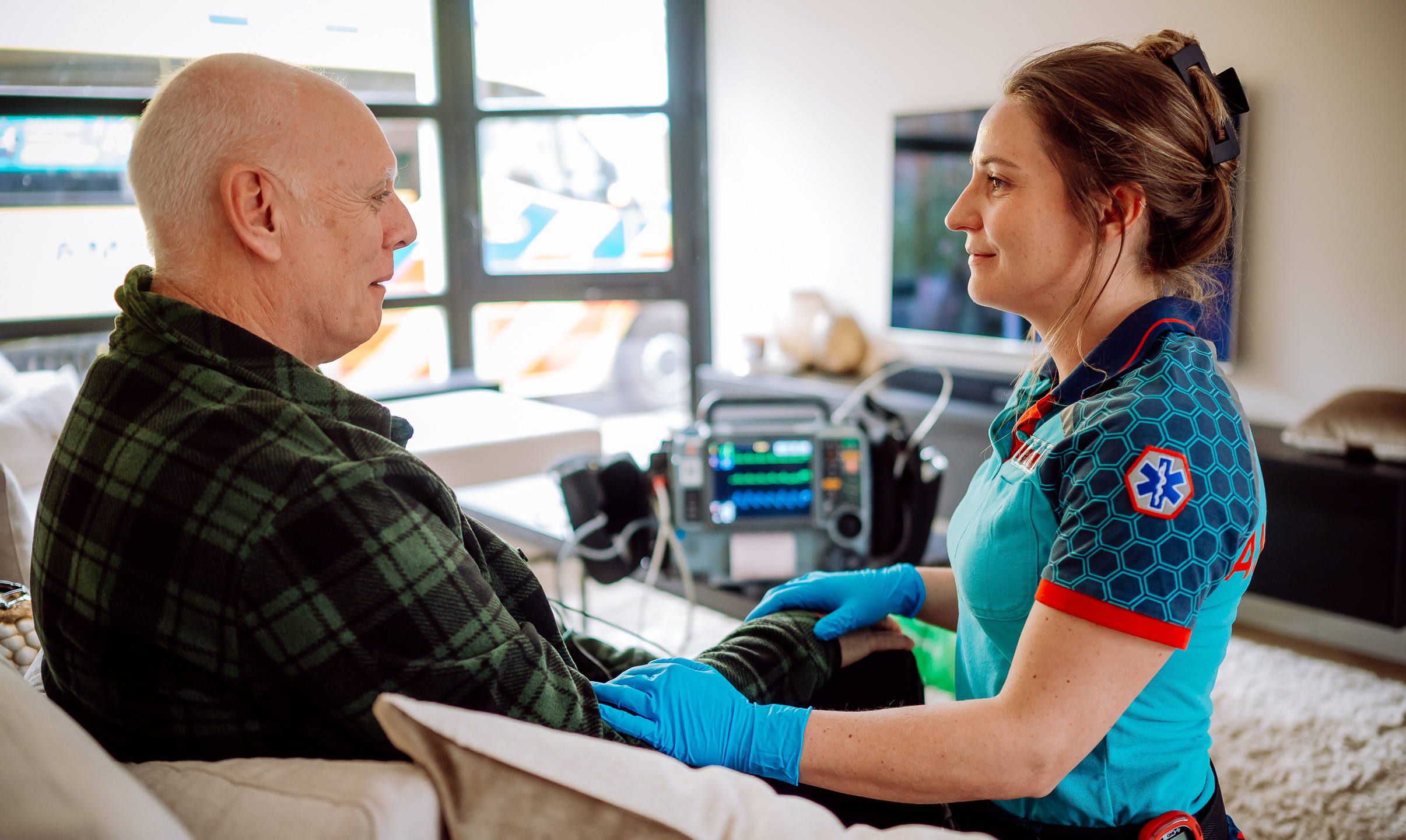 Ambulanceverpleegkundige helpt een patiënt in de woonkamer. Er ligt een monitor op de achtergrond