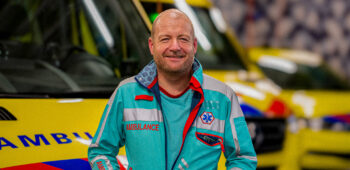 Ambulanceverpleegkundige Jorre kijkt lachend in de camera met ambulances op de achtergrond