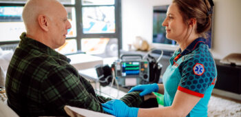 Ambulanceverpleegkundige helpt een patiënt in de woonkamer. Er ligt een monitor op de achtergrond