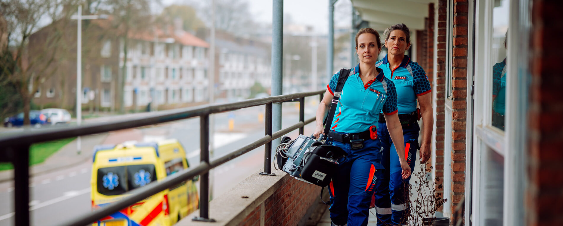 Een ambulanceteam loopt over de galerij van een appartement richting de hulpverlening. De ambulanceverpleegkundige heeft een monitor in haar hand. Op de achtergrond is de ambulance te zien die geparkeerd staat.