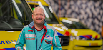 Ambulanceverpleegkundige Jorre kijkt lachend. Op de achtergrond is een ambulancehal waarin ambulances geparkeerd staan.