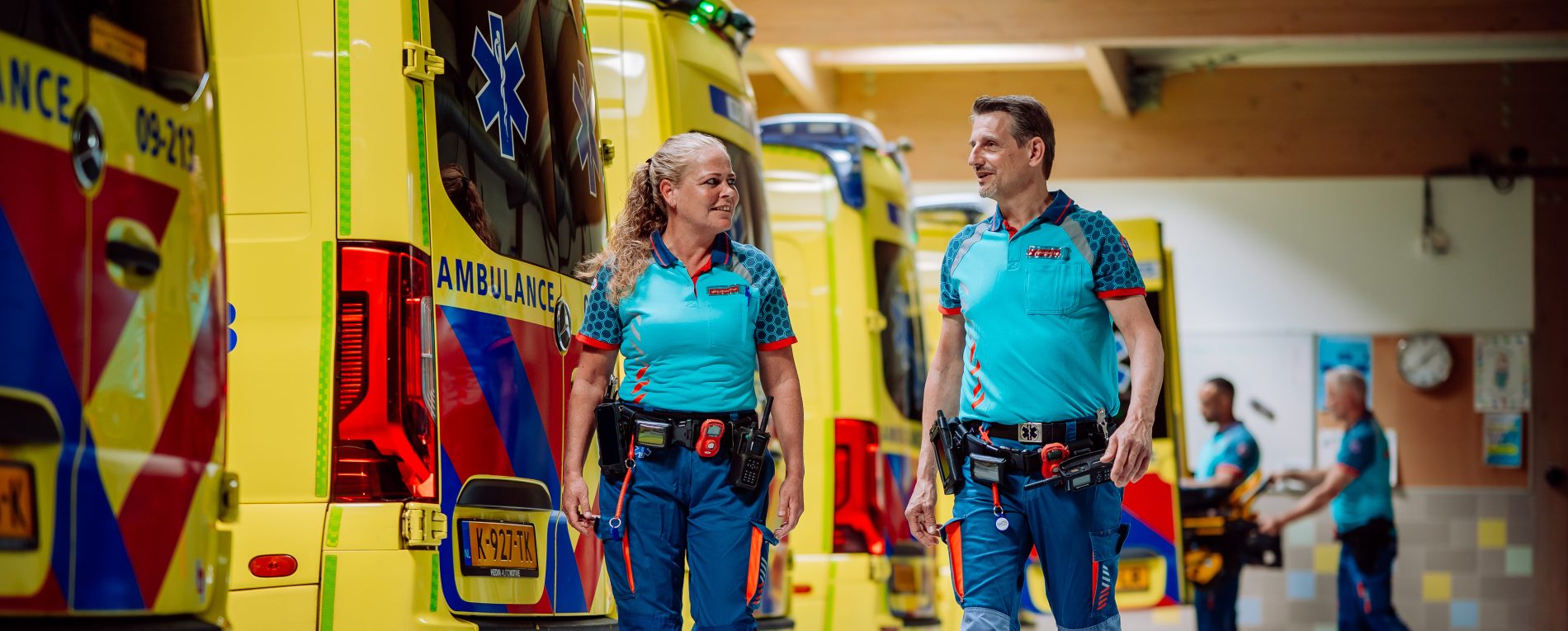 Een ambulanceteam loopt door de ambulancehal. Ze kijken elkaar aan en lachen. Op de achtergrond is een ander ambulanceteam bezig met een brancard.